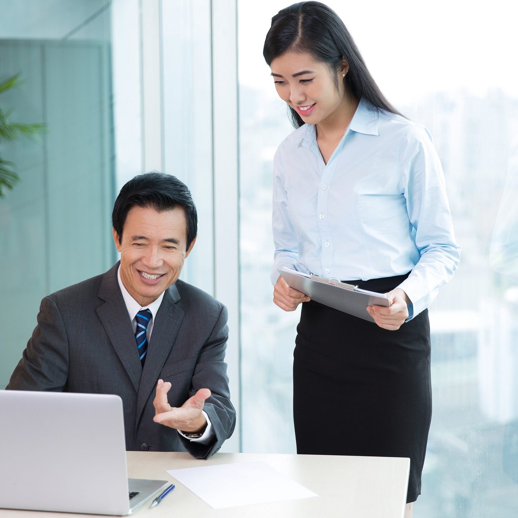 Smiling Asian senior business man pointing to laptop screen and sitting at desk while talking to female assistant standing near in office with window and blurred city view outside in background